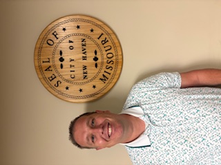 A man standing indoors beside a circular wall seal that reads “SEAL OF CITY OF NEW HAVEN, MISSOURI.”